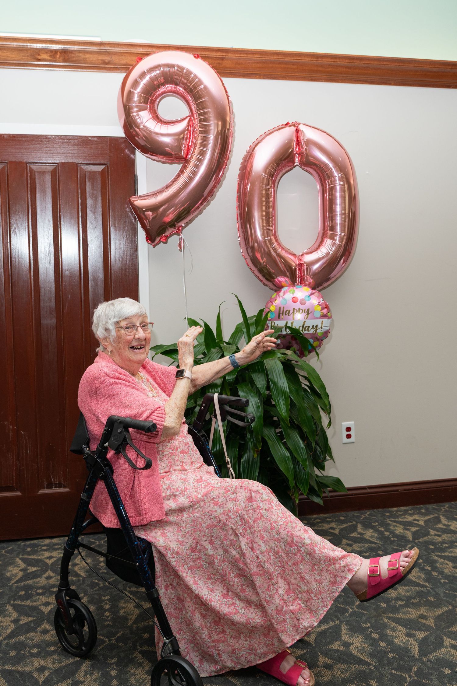 Elderly woman in pink dress celebrates 90th birthday with rose gold balloons and happy birthday balloon.