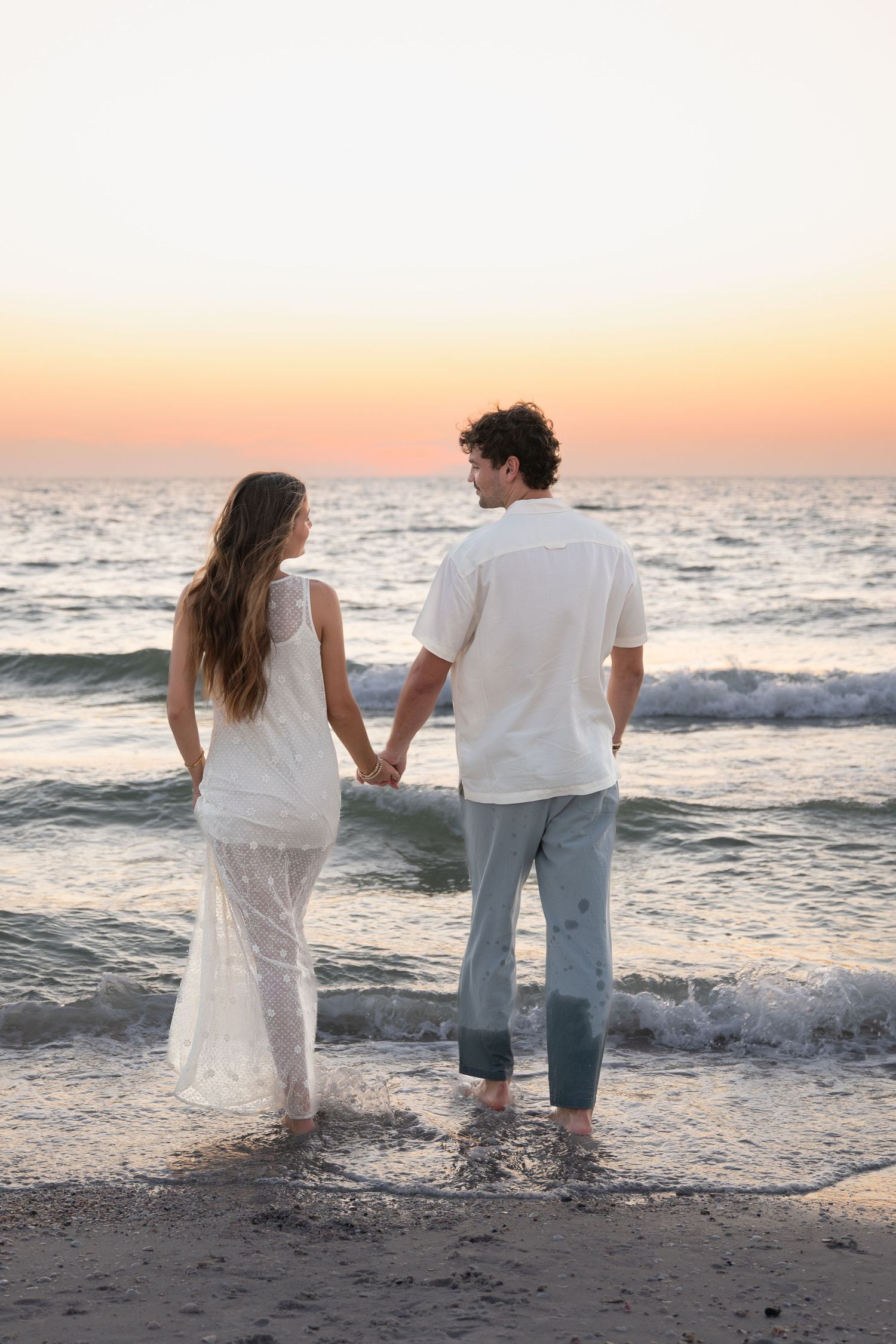 Couple walking hand in hand in shallow ocean waves at sunset.