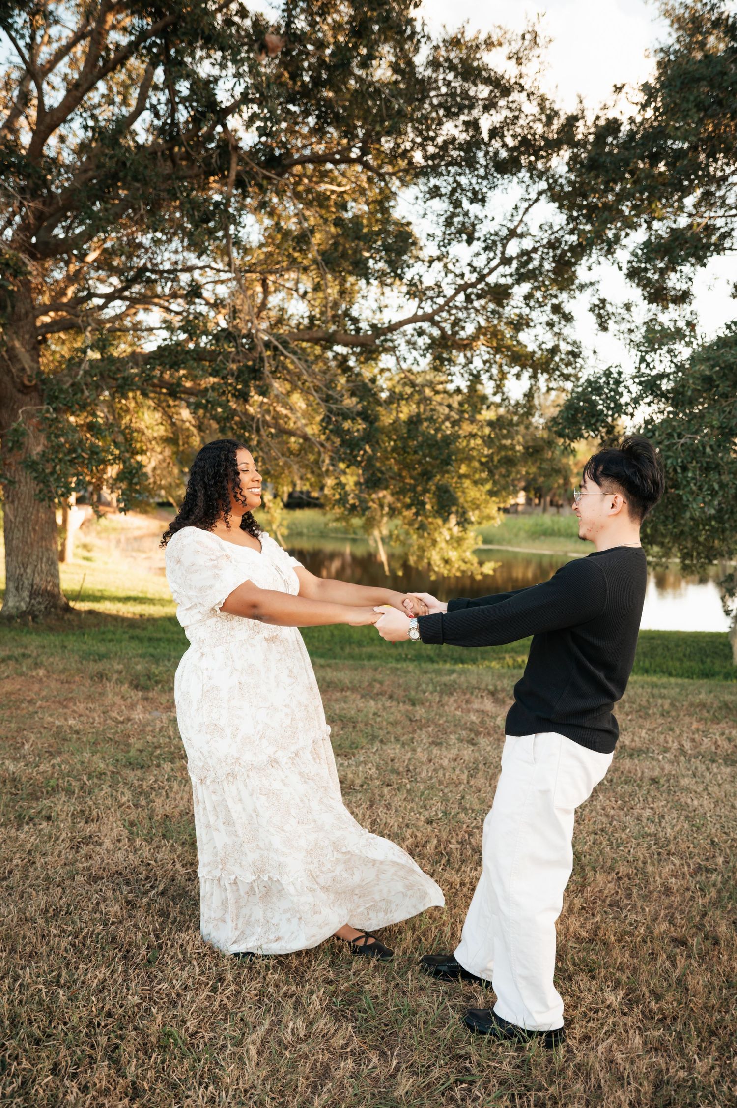 A couple in white and black attire dance playfully in a sunlit field during golden hour.
