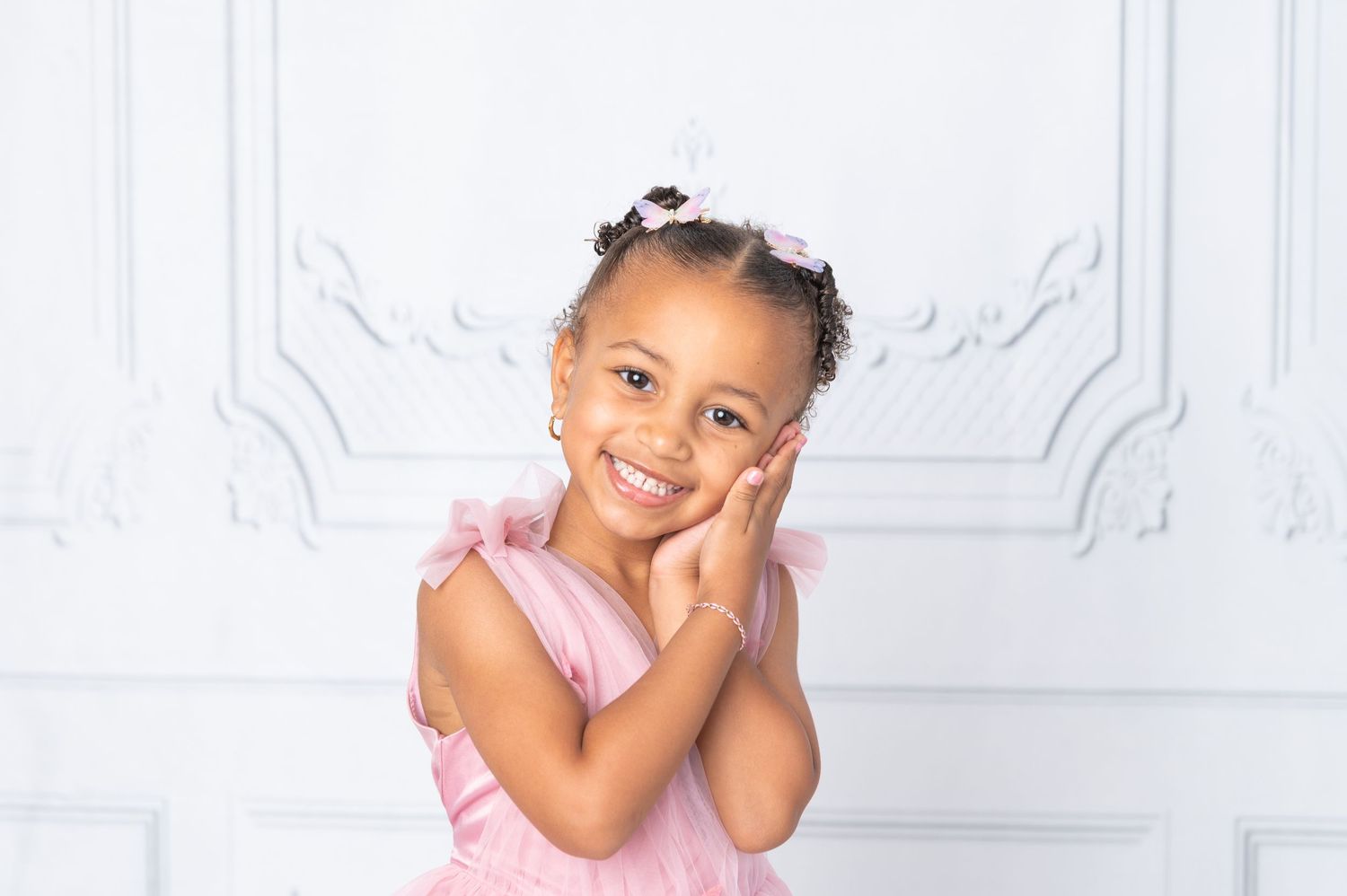 A smiling young girl in a pink dress poses with hands on cheeks against a white ornate wall backdrop.