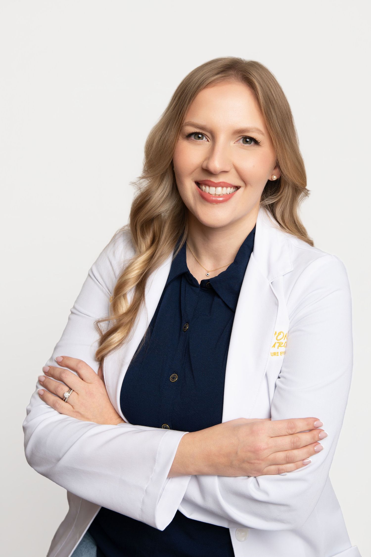 Smiling female healthcare professional in white lab coat with arms crossed against a light gray background.