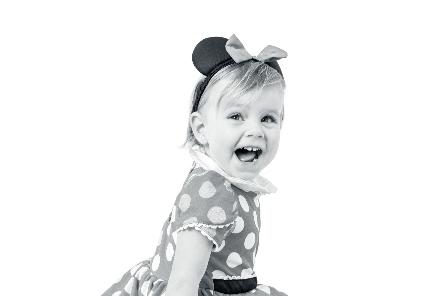 Laughing toddler girl in Minnie Mouse polka dot dress and ear headband, black and white portrait photo.