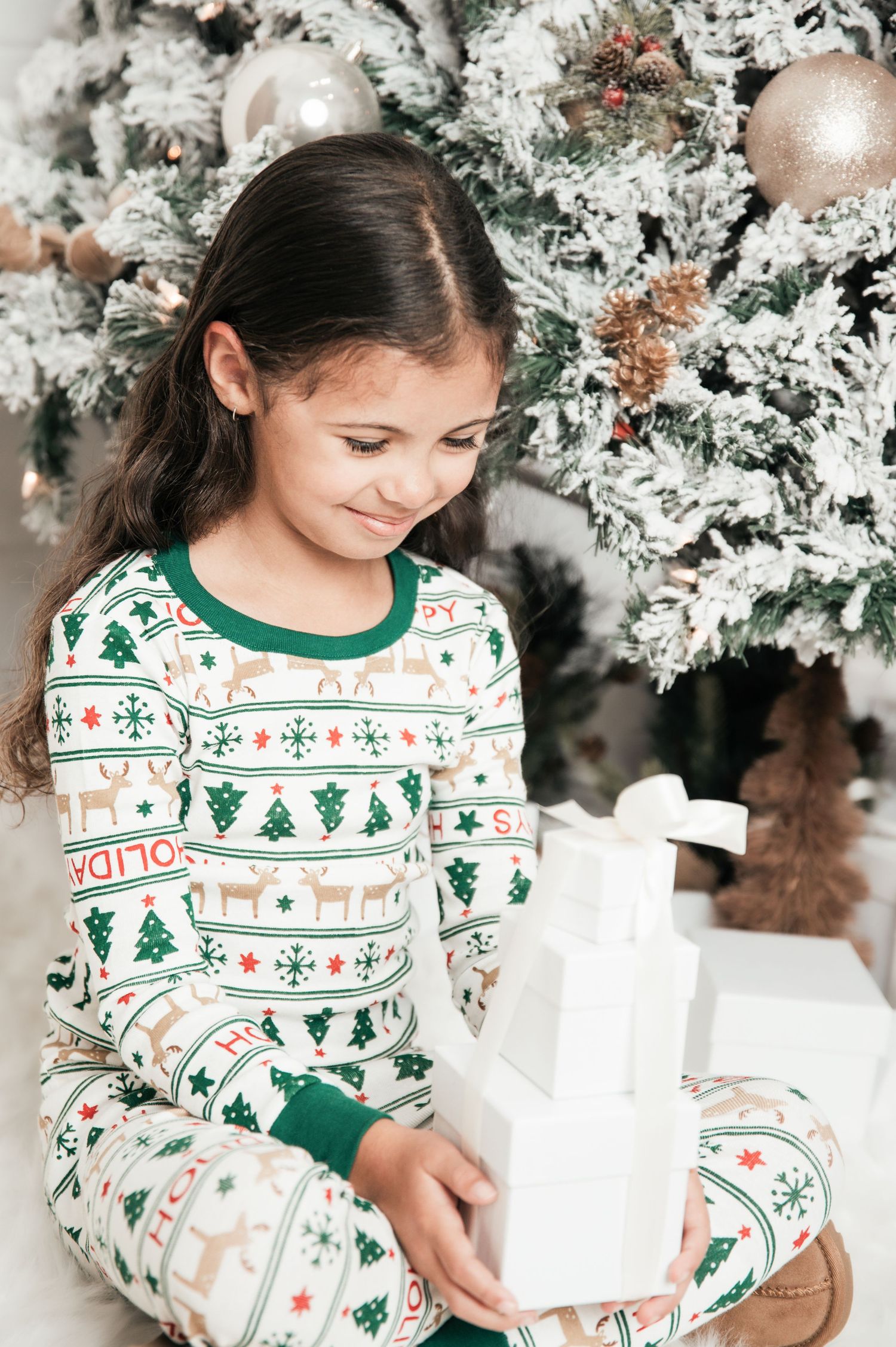 Child in festive Christmas pajamas with green and white pattern sits by decorated white flocked tree with holiday ornaments.