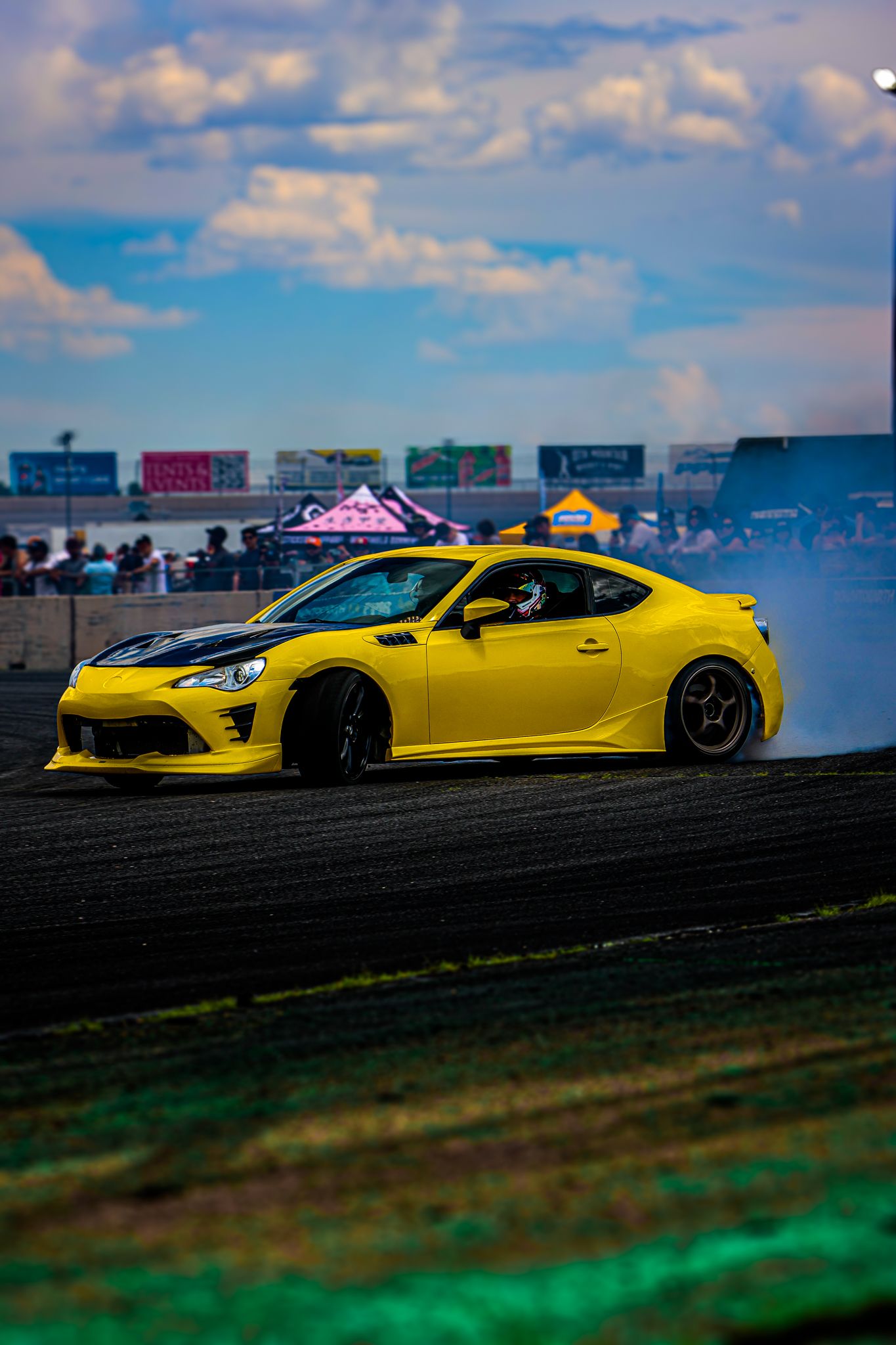 A bright yellow sports car sits parked on a grassy hillside against a blue sky with white clouds.