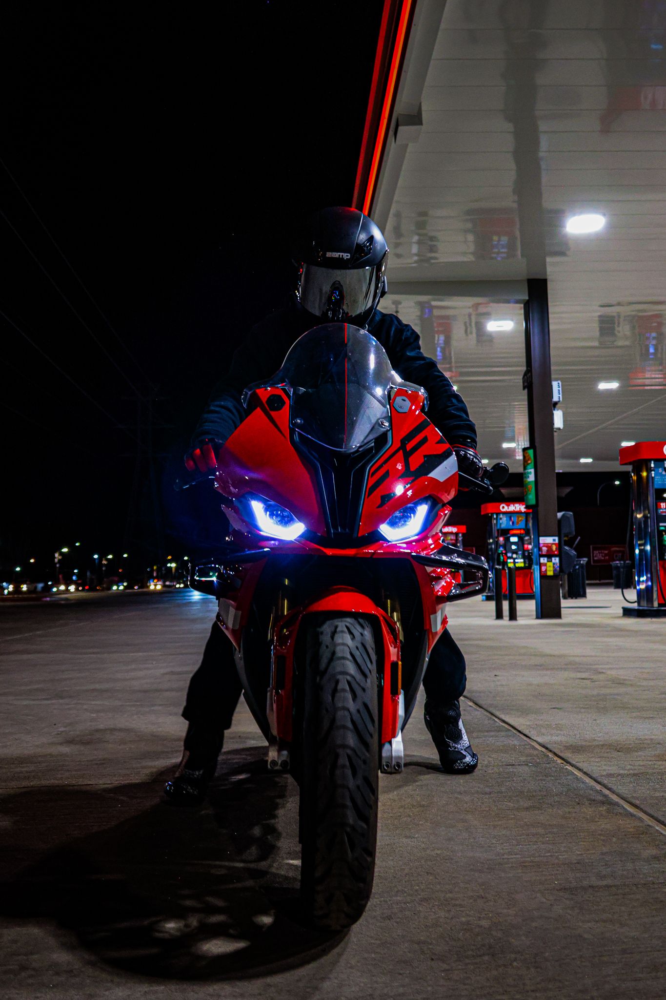 Red sport motorcycle with blue LED headlights at night gas station, rider in black helmet and gear standing behind bike.