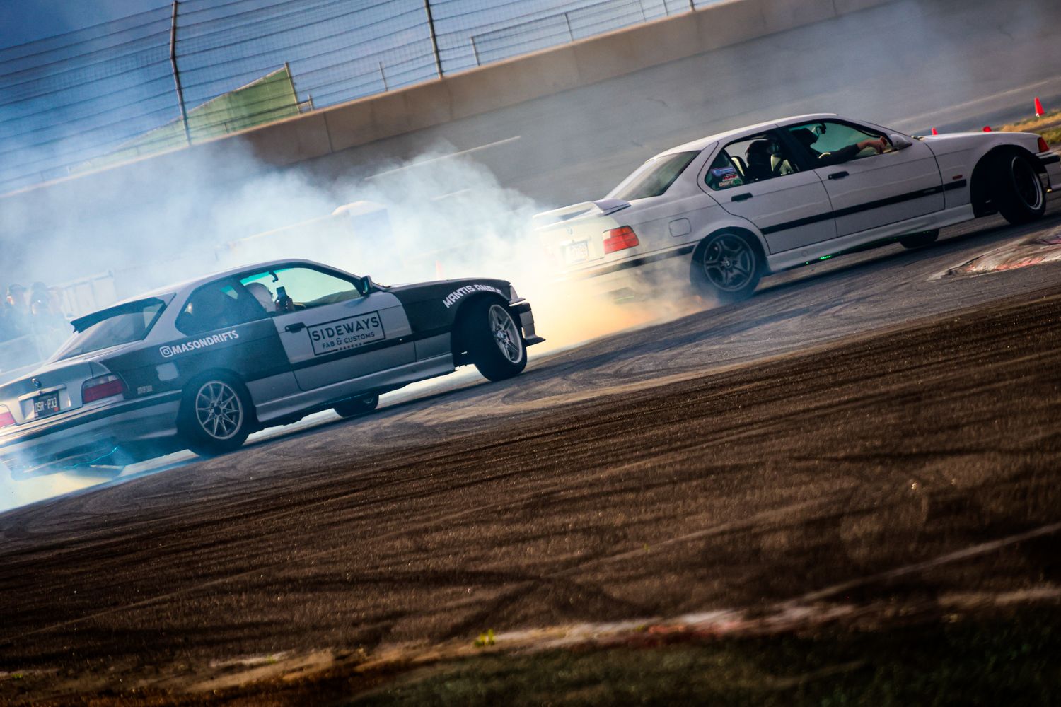 Two BMW cars drifting on a dirt track, creating large smoke clouds during an exciting motorsport event.