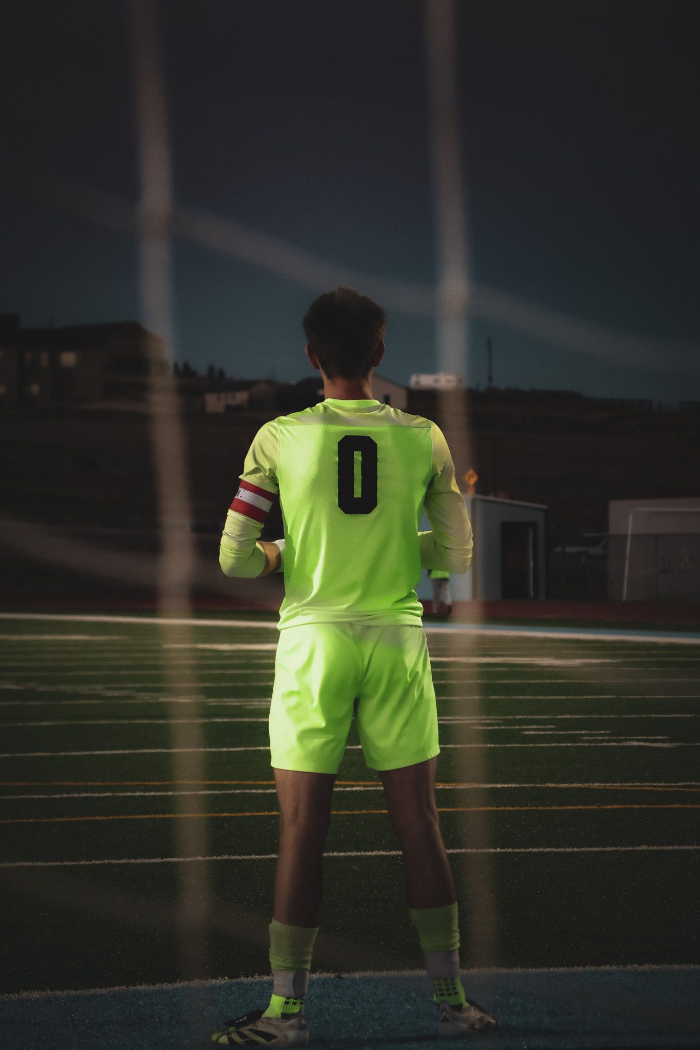 Soccer player wearing neon yellow jersey with number 0 stands on field at night under stadium lights.
