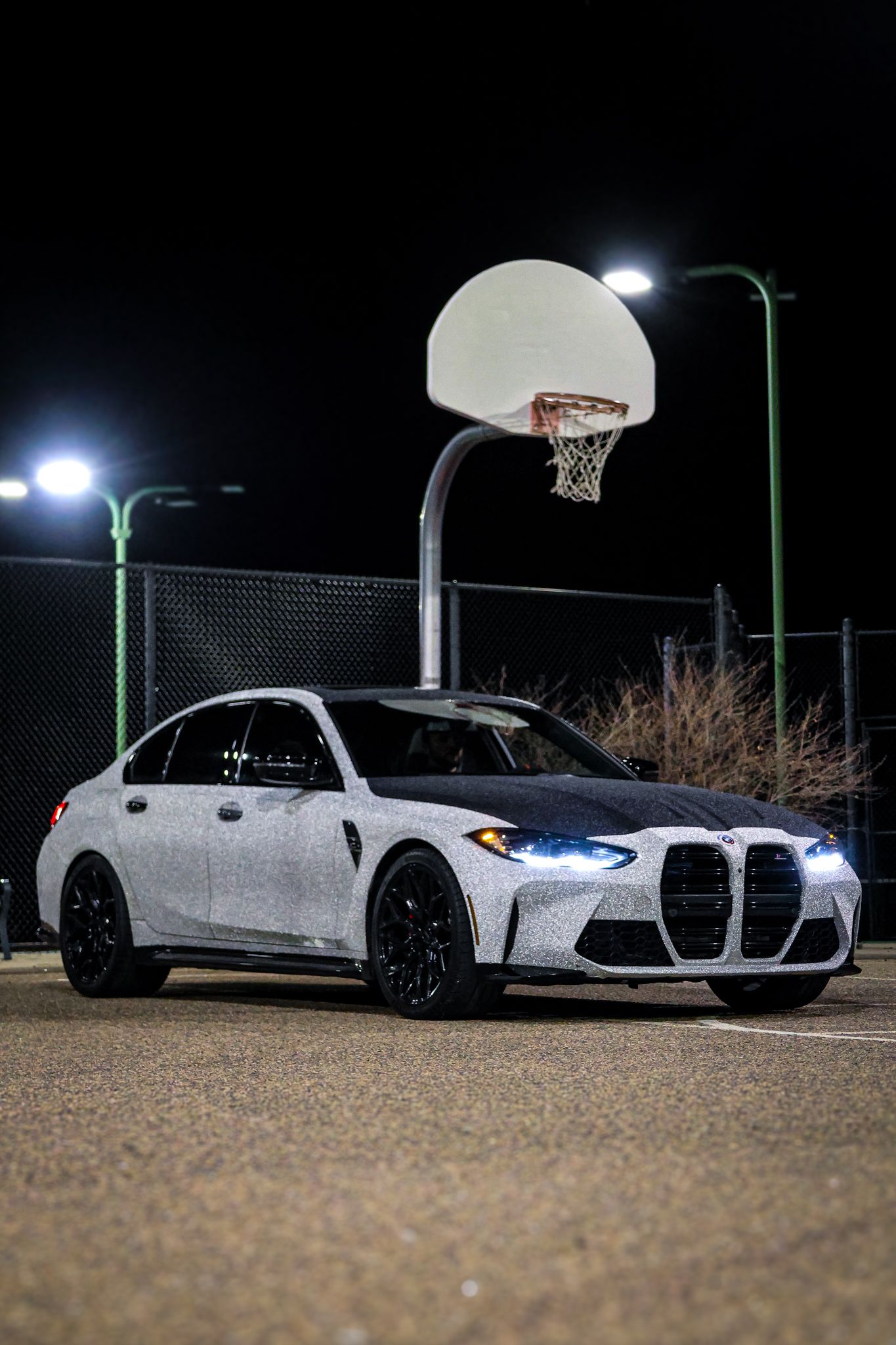A white BMW M3 parked at night under a basketball hoop with illuminated headlights.