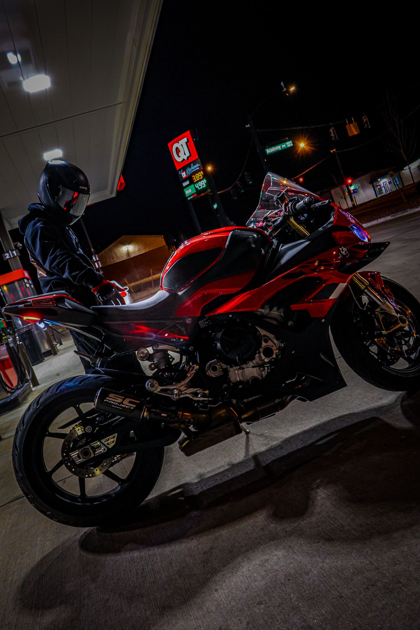 Red sport motorcycle parked at night gas station with helmeted rider, dramatic dark lighting highlighting chrome and bodywork details.