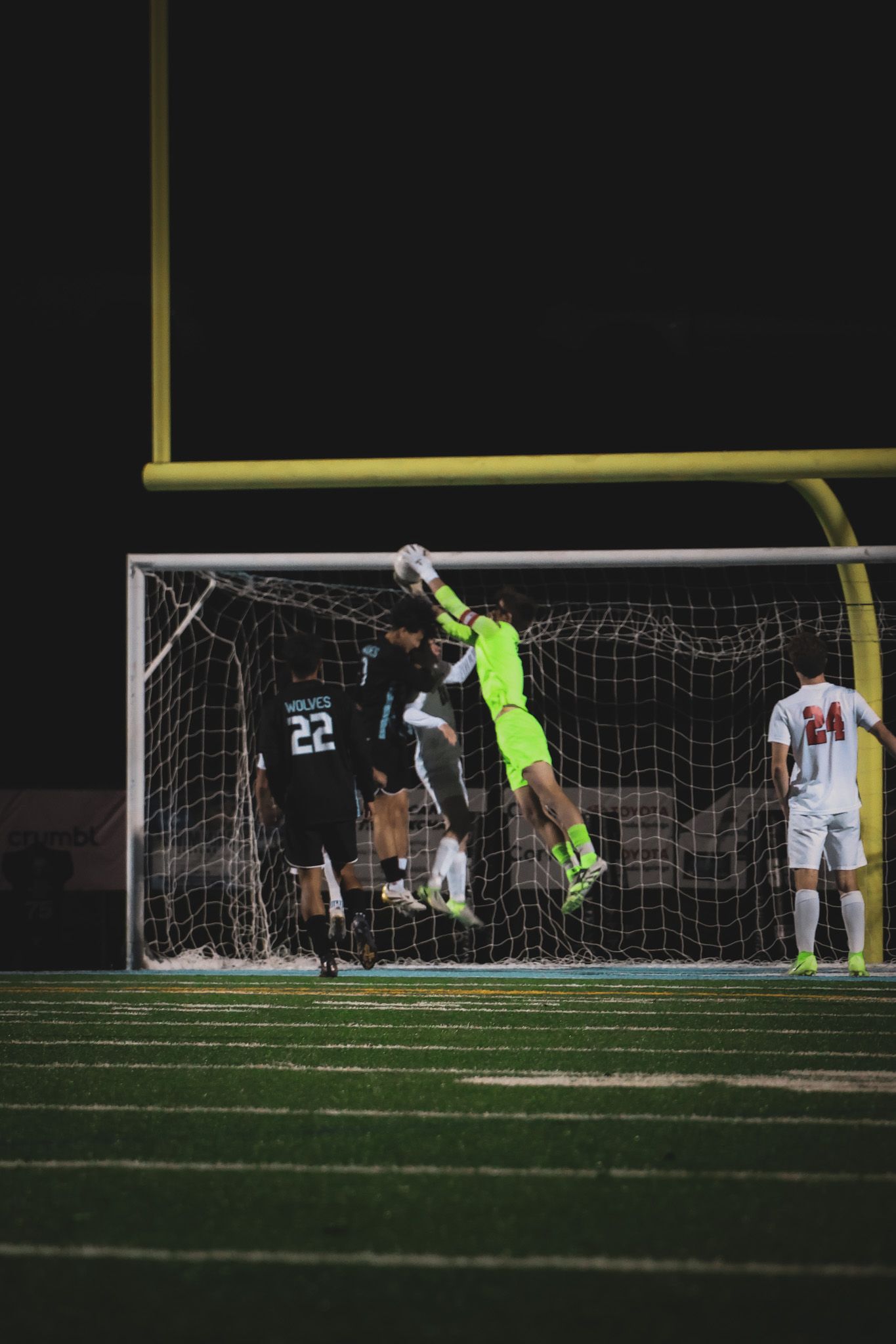 Soccer goalkeeper in bright yellow jersey makes diving save attempt at goal during nighttime match on turf field.