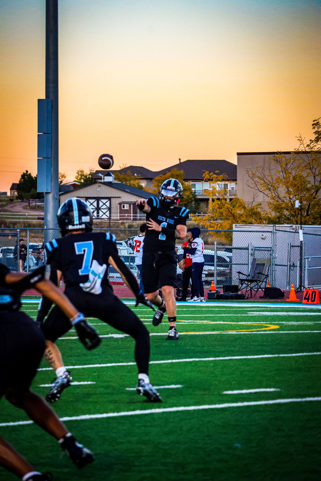 Football players in black uniforms compete on an artificial turf field during a sunset game with stadium lights illuminating.