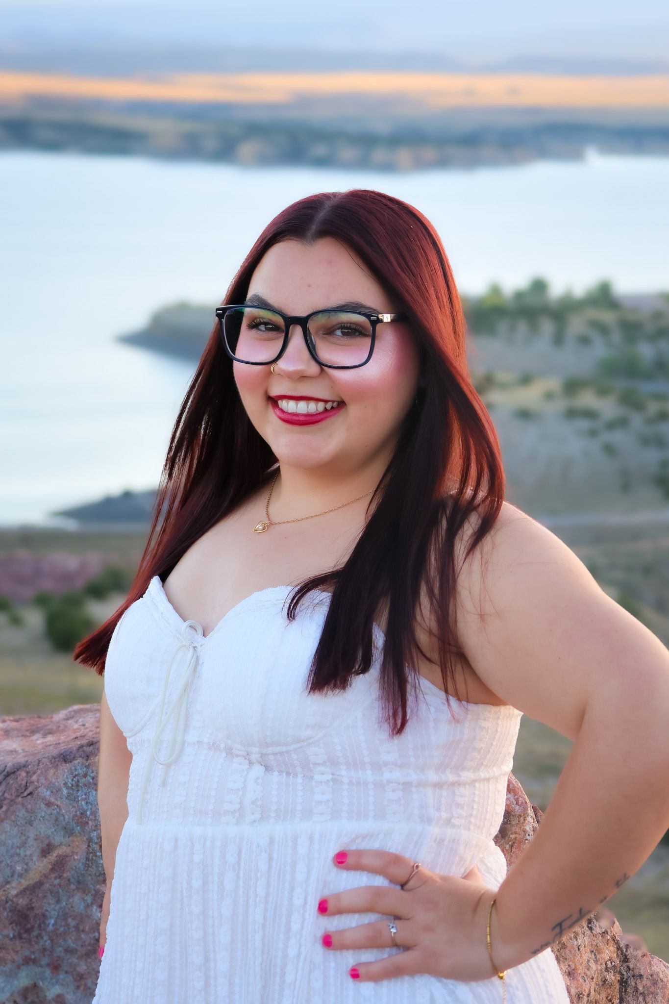 A smiling person in glasses and a white dress poses outdoors with a scenic lake and sunset view in the background.