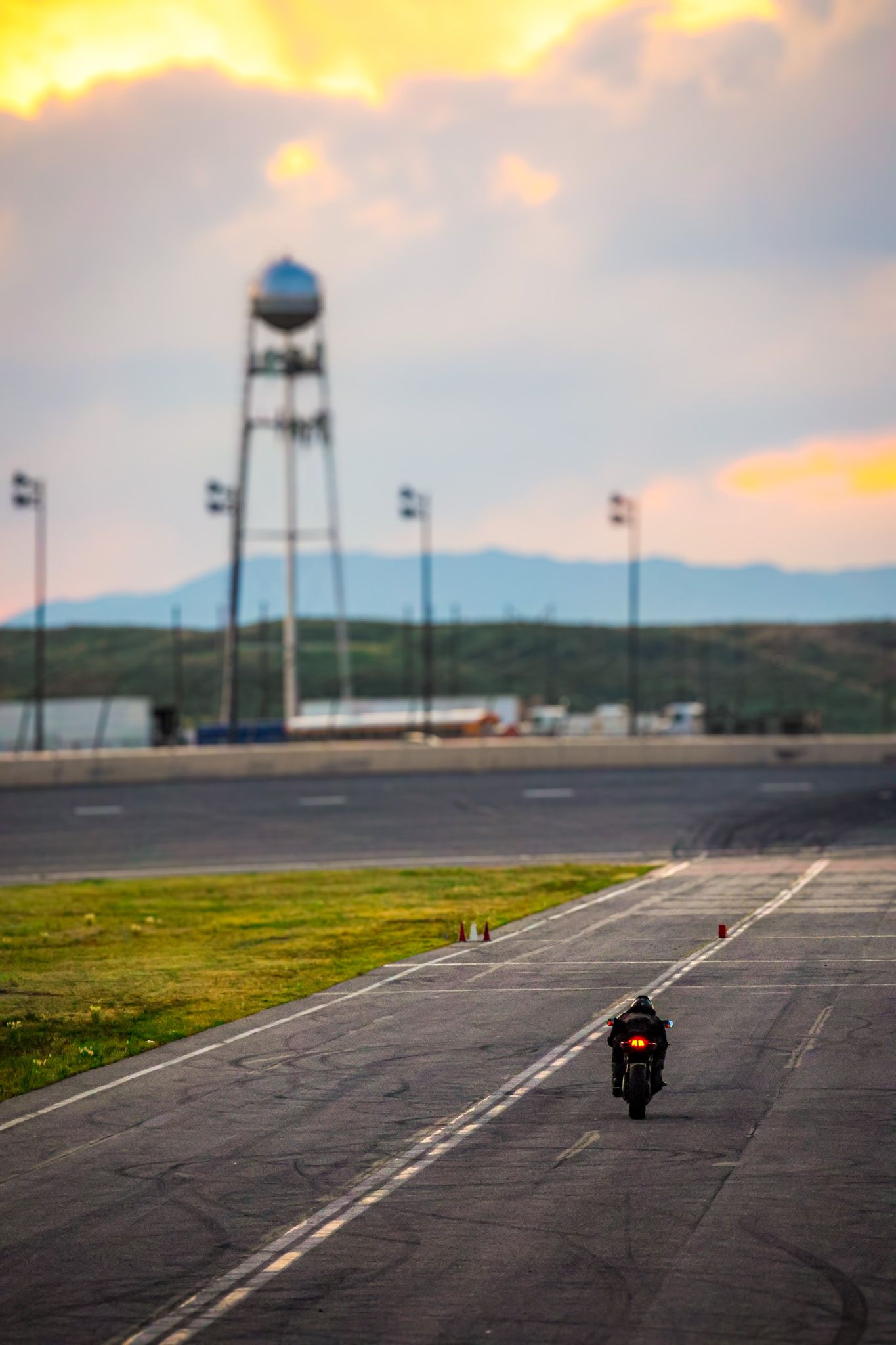 Motorcycle rider on empty racetrack with water tower silhouetted against dramatic sunset sky.