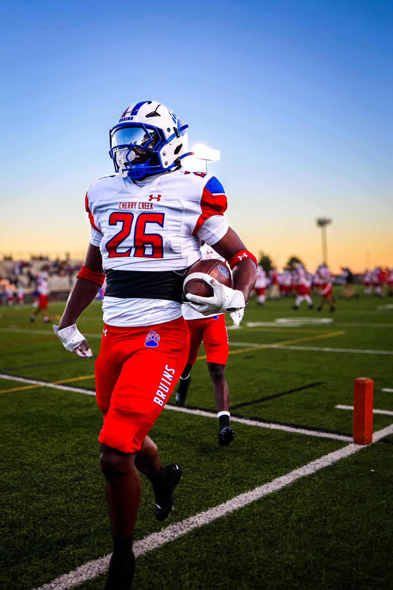Football player in white and red uniform runs drills during sunset practice on green turf field.