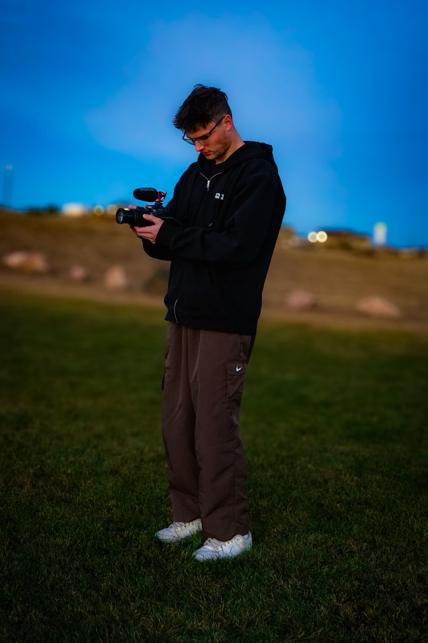 A photographer in dark clothing stands on grass against a bright blue sky taking photos with a digital camera.