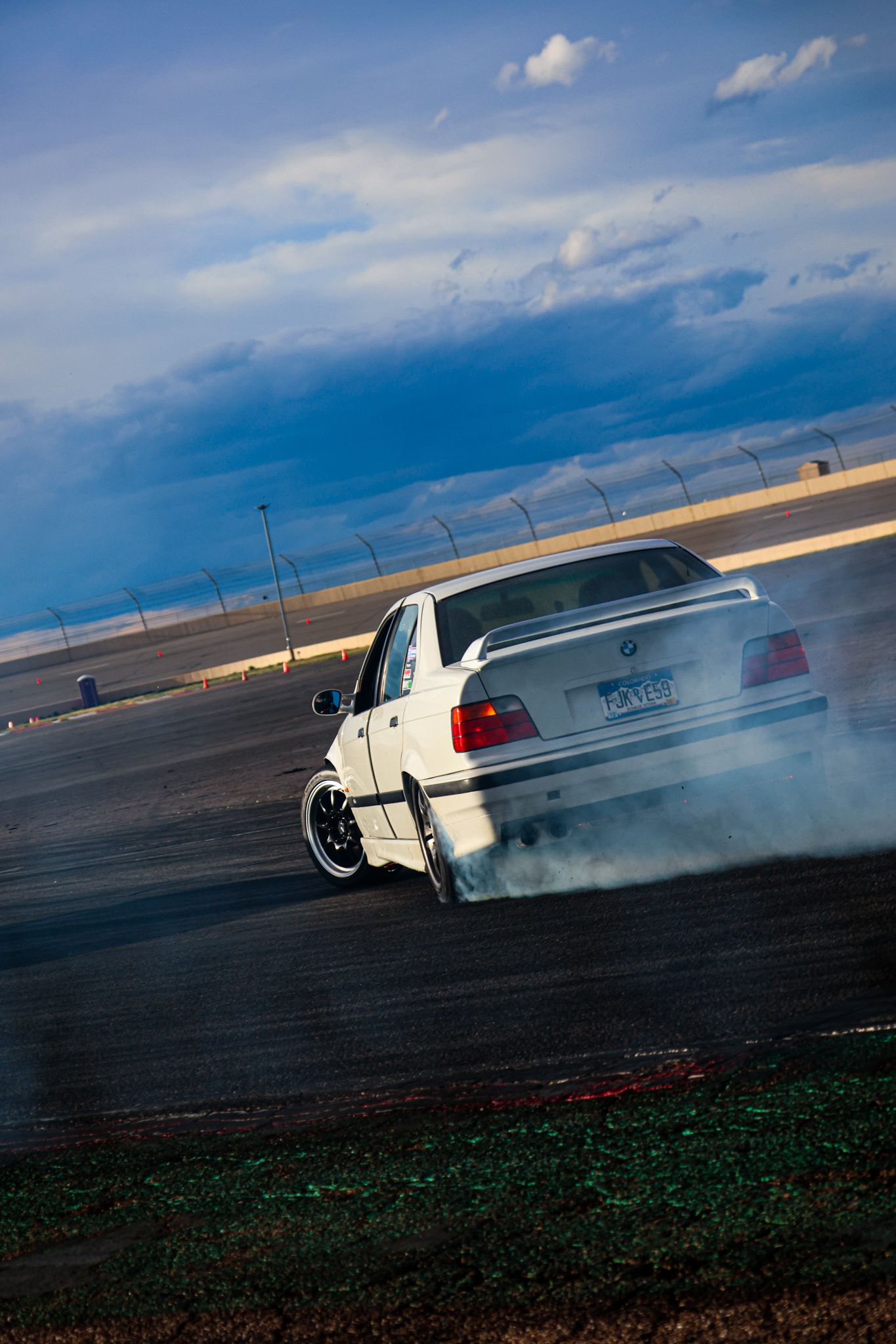 White BMW drifting on a racetrack, producing tire smoke under a dramatic blue cloudy sky.