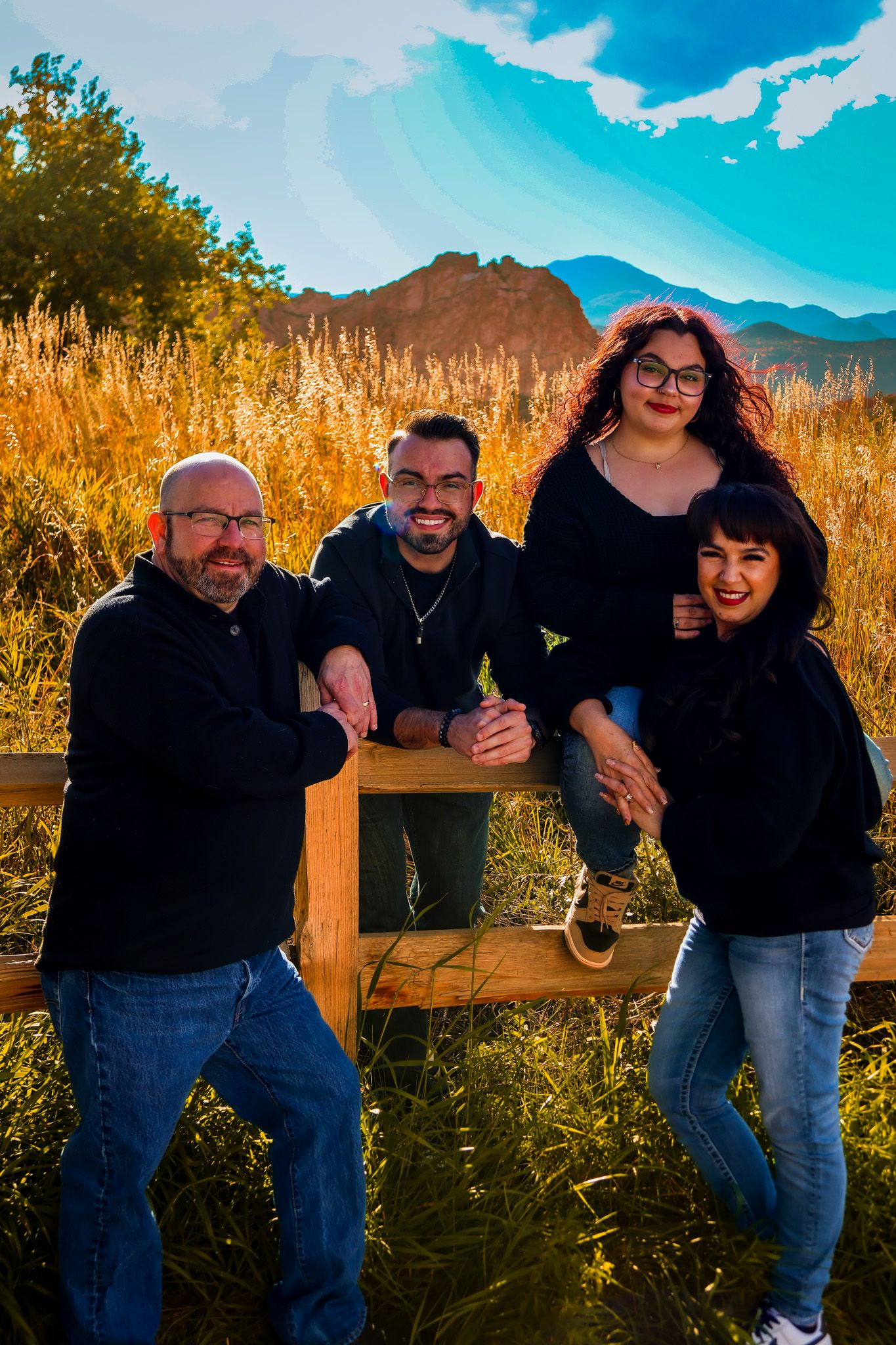A group poses playfully on a wooden fence with mountains and autumn foliage in the background during golden hour.