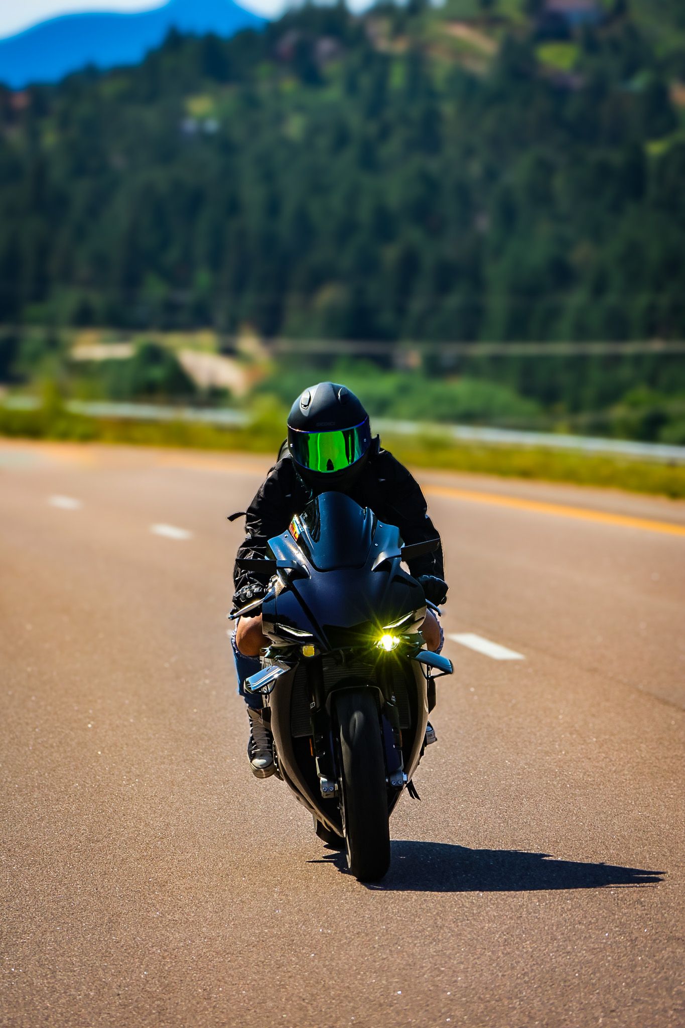 A motorcyclist in black gear rides a sports bike on an empty mountain road with evergreen trees in the background.