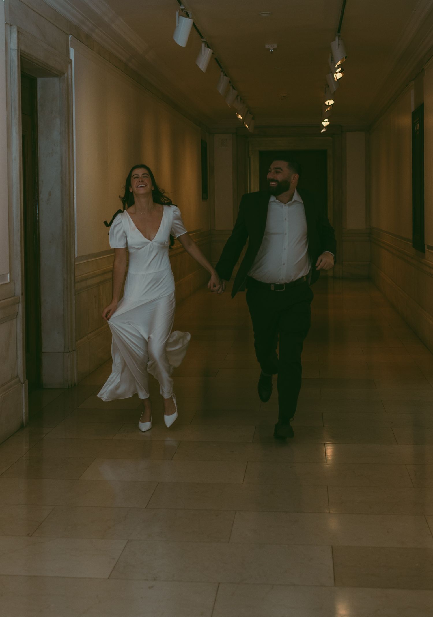 A couple runs playfully through an elegant hallway, with the woman in a white dress and man in dark suit.