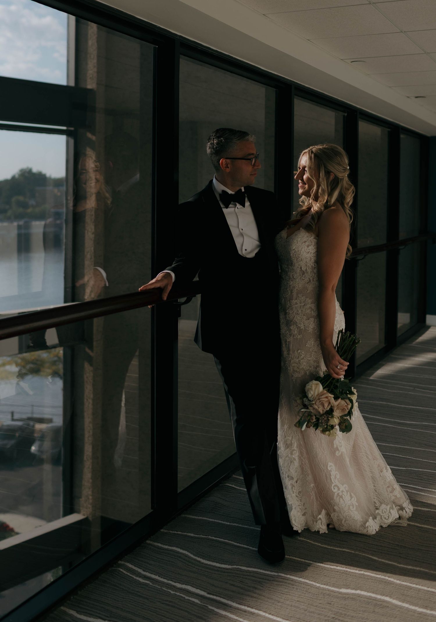 Couple in formal attire poses by large windows with city views.