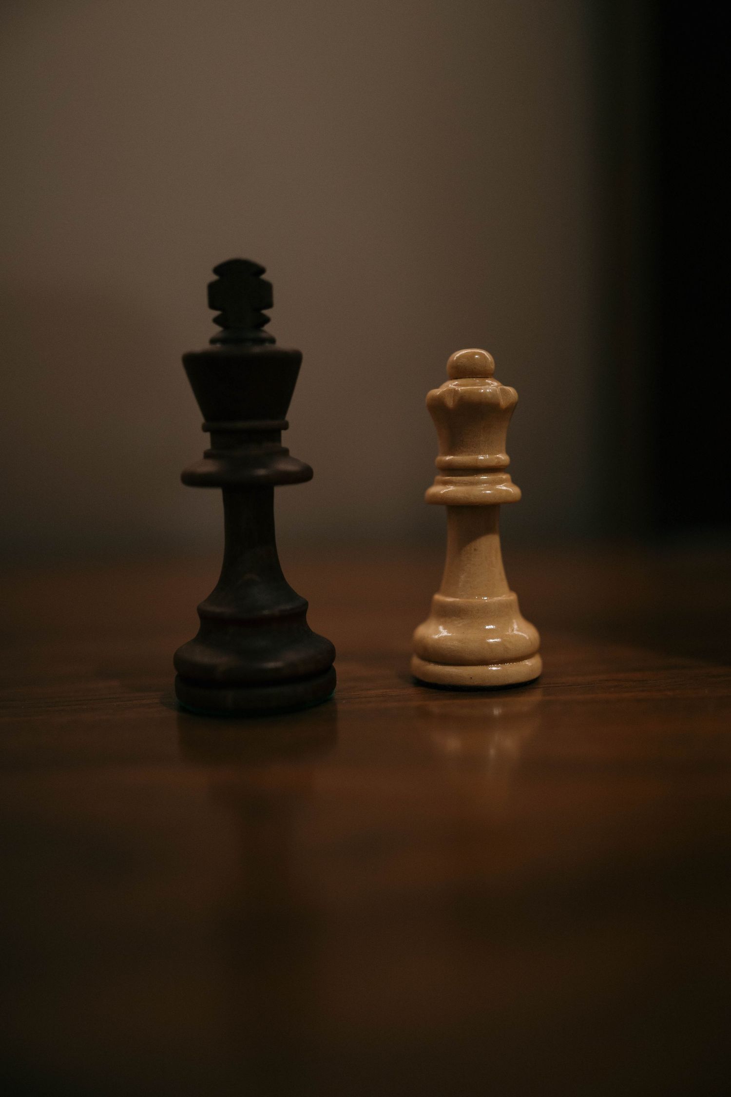 A black king and white queen chess piece stand together on a dark wooden surface in dramatic lighting.