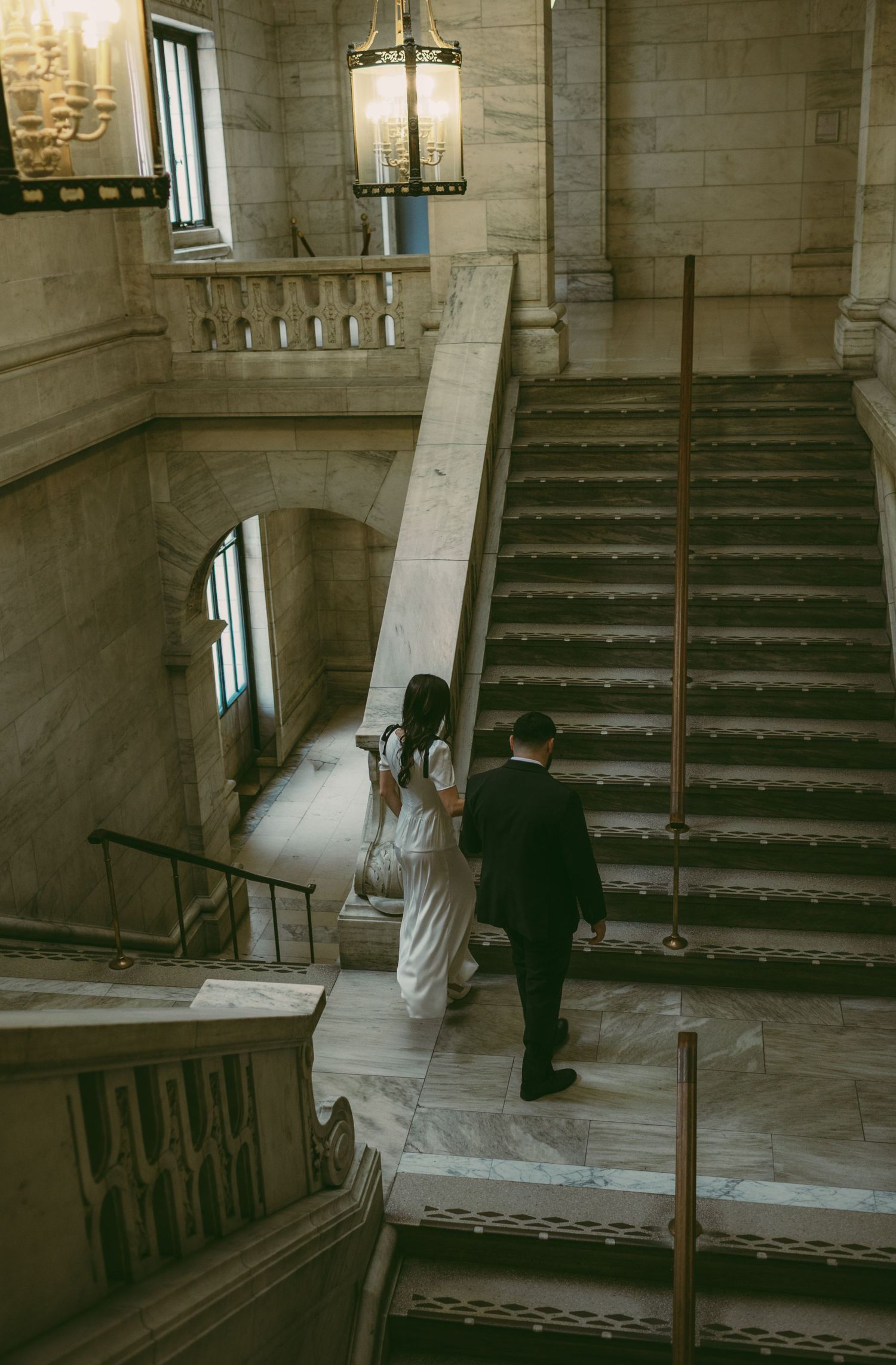 Two people ascend a grand marble staircase with vintage lighting fixtures.