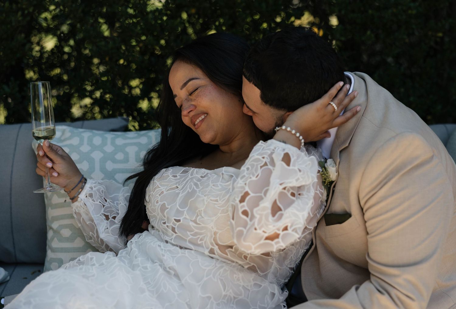 A couple shares an intimate moment on a couch, with the bride wearing a white lace dress and holding a champagne glass.