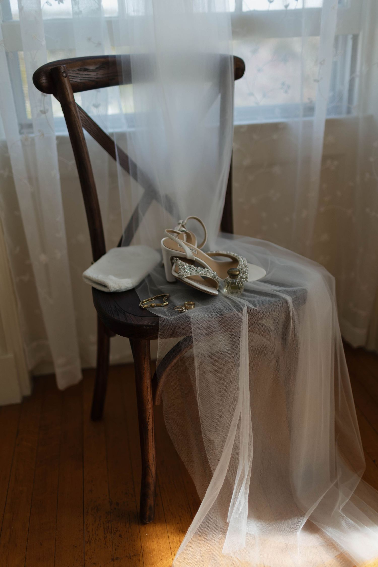 Wedding shoes and veil draped elegantly over a vintage wooden chair near sheer curtains.