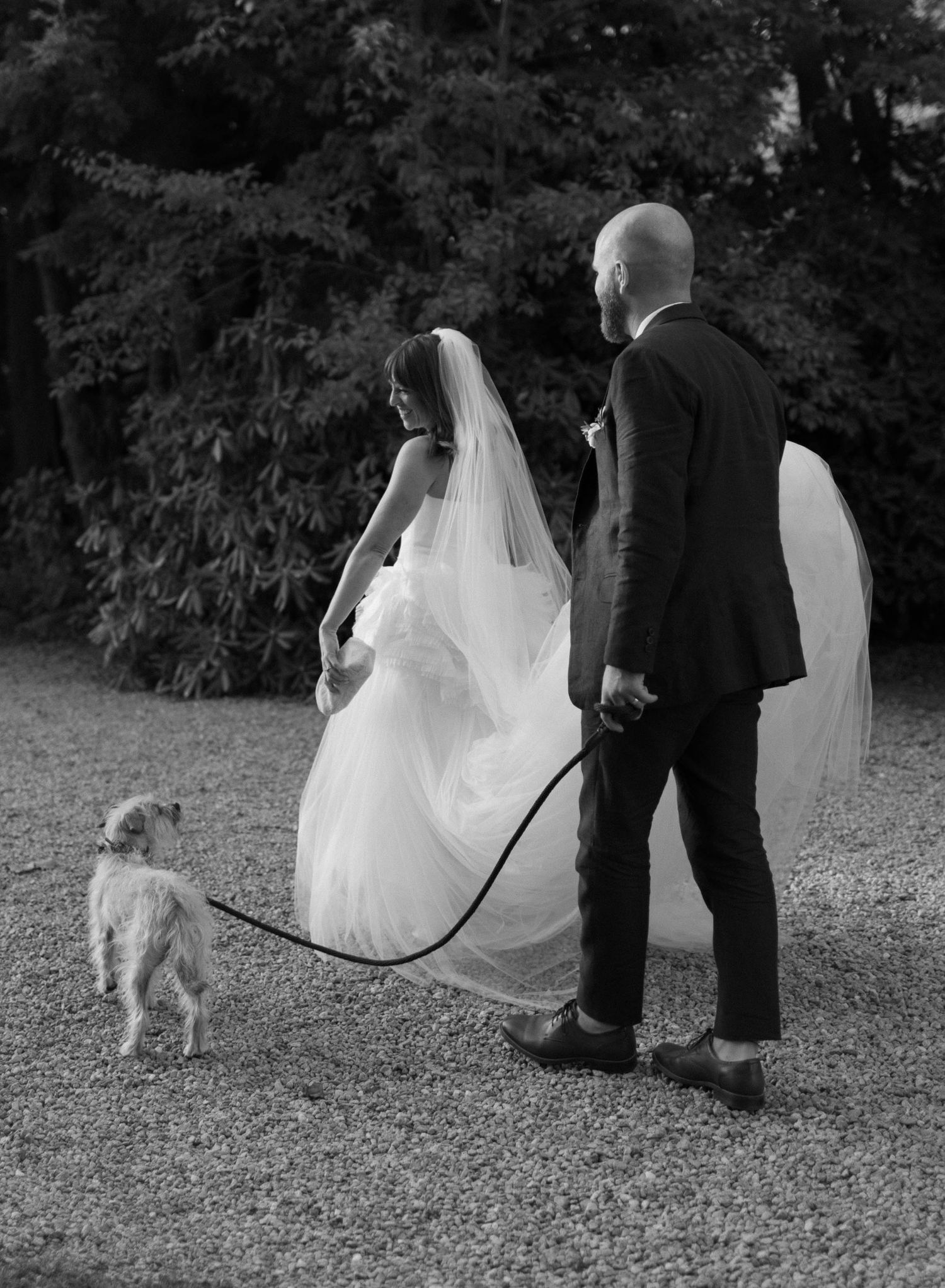 Black and white photo of couple walking with dog on gravel path.