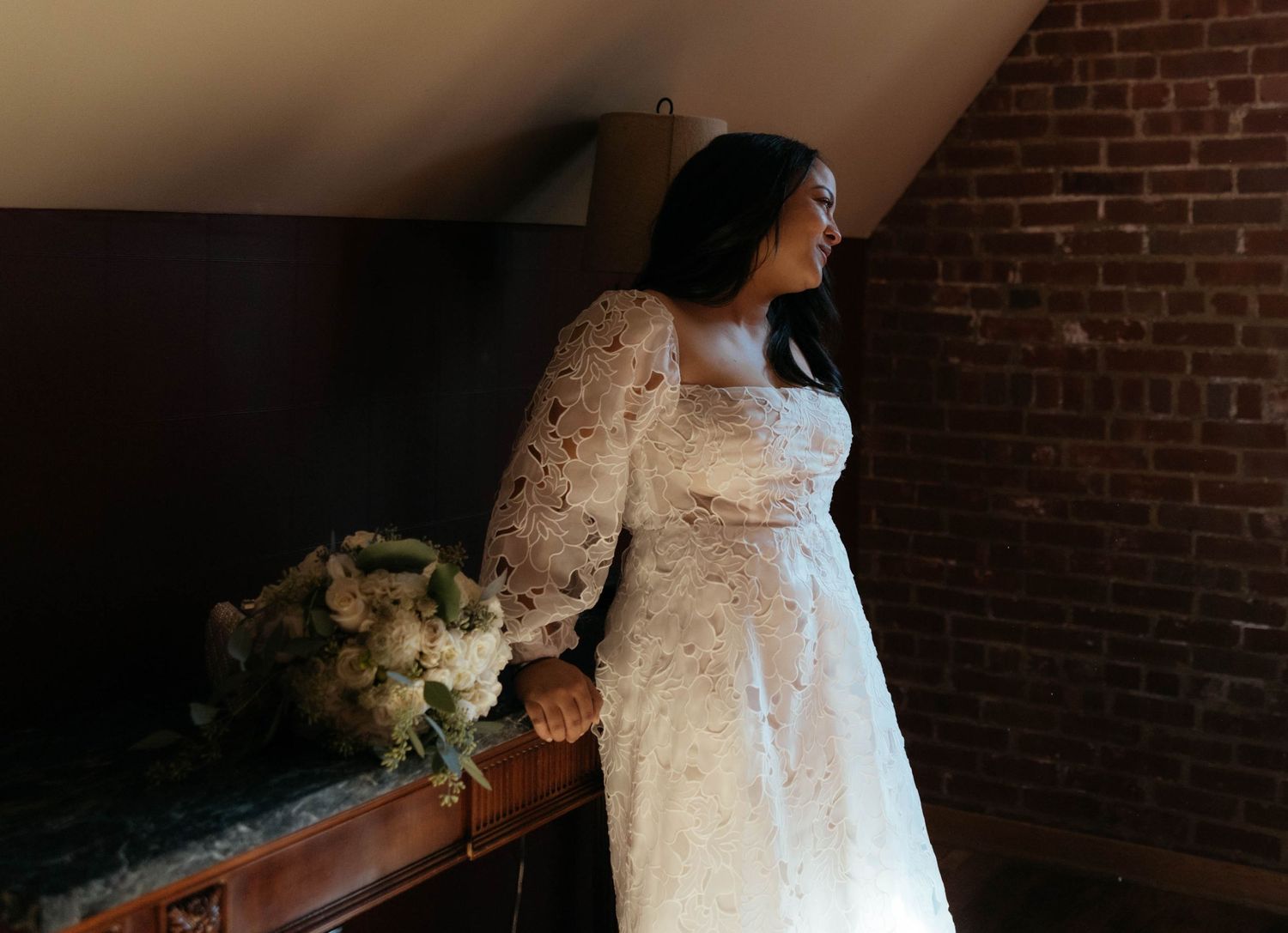 White lace wedding dress detail in front of exposed brick wall and window.