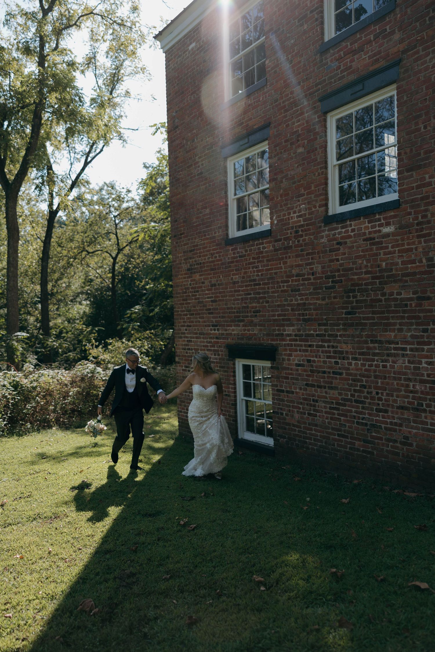 Newlyweds walking together by a sunlit brick building with white-trimmed windows.