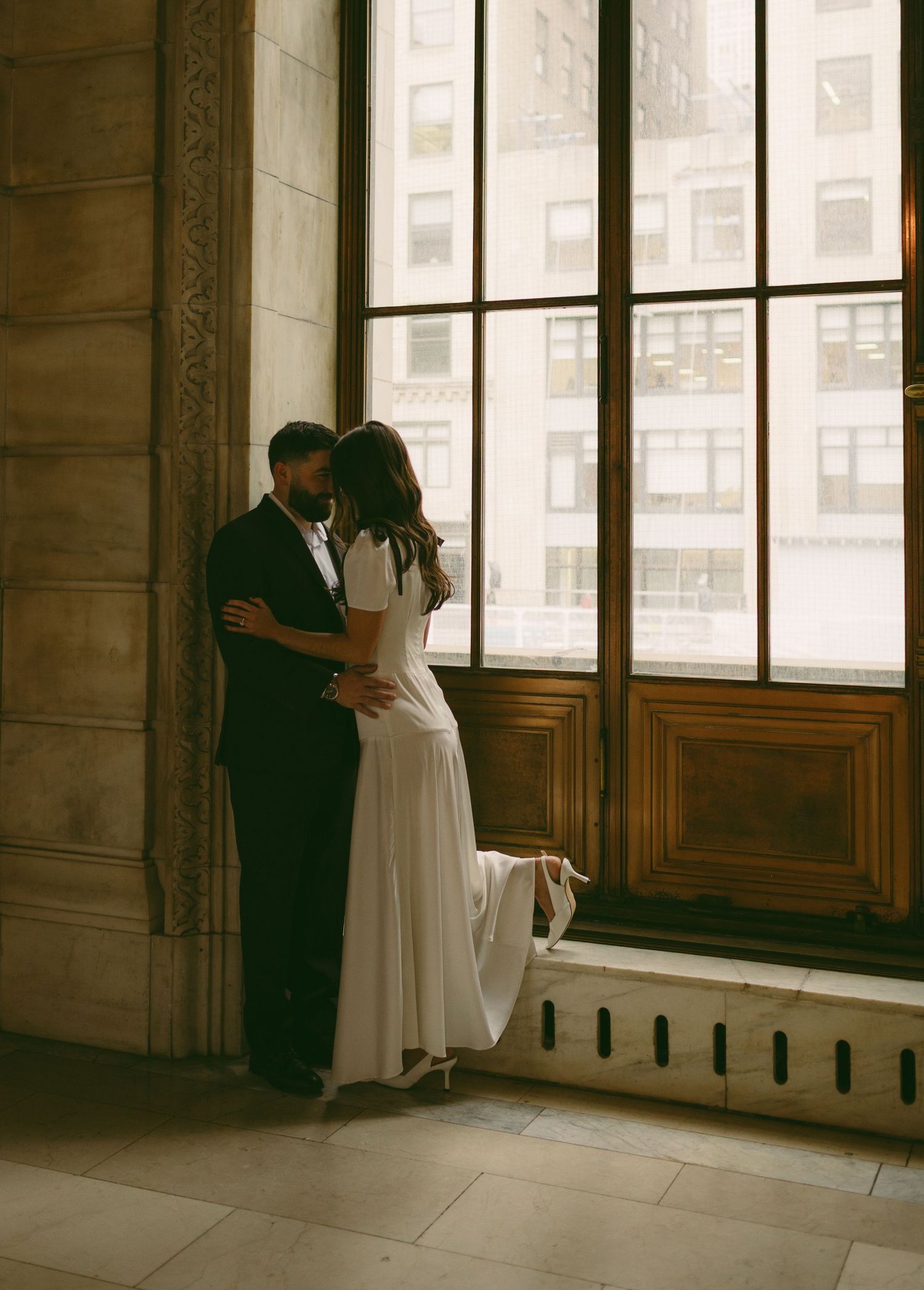 Couple in formal attire embracing by large windows in an elegant interior.