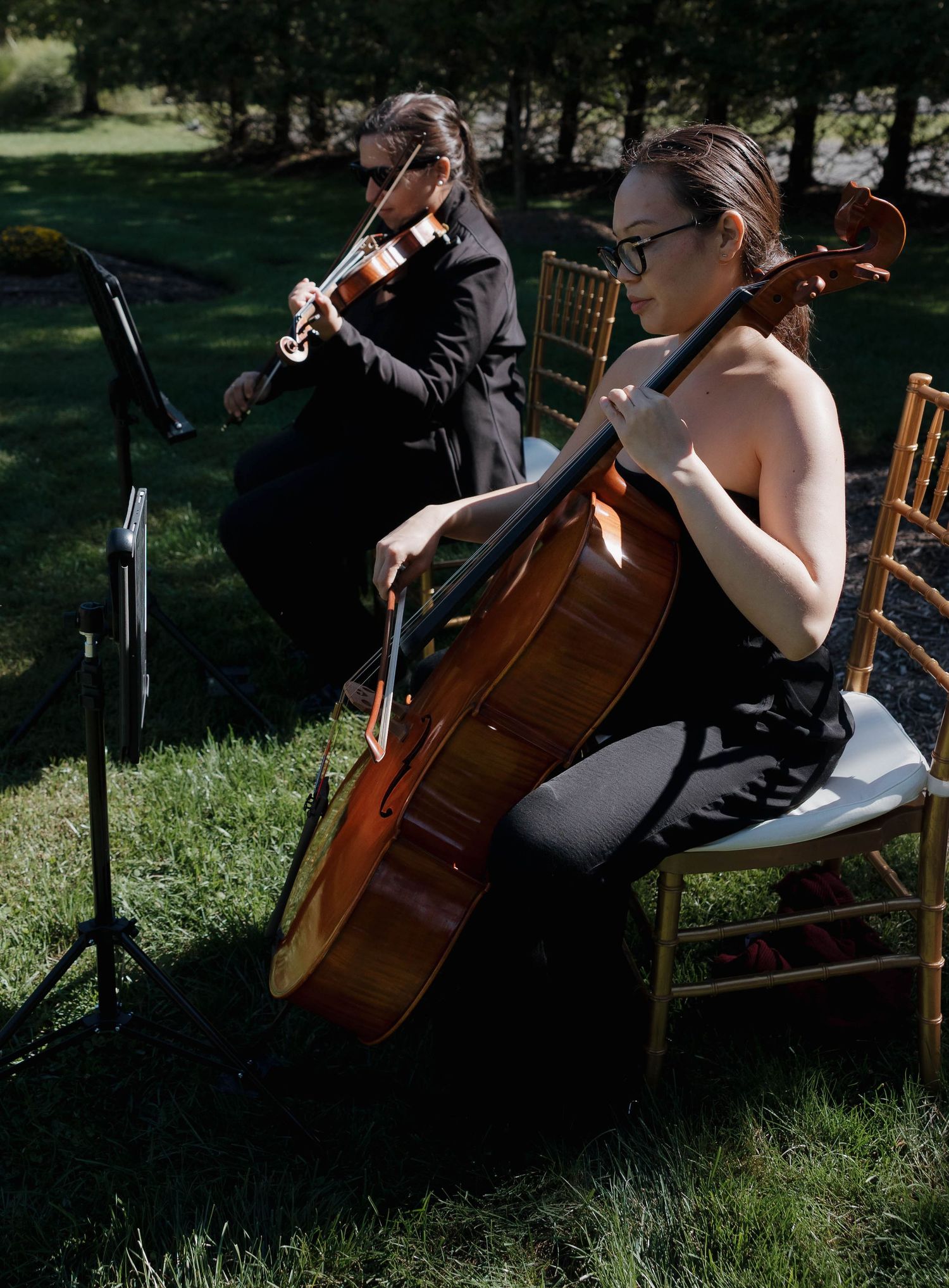 Musicians play cellos outdoors on wooden chairs during a wedding ceremony.