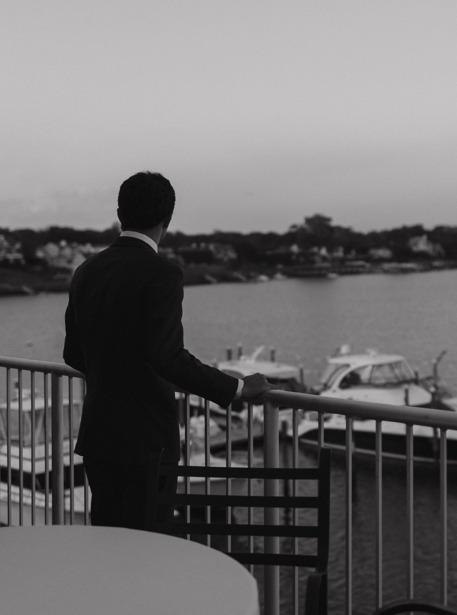 A silhouetted figure in a suit gazes out over a marina from a balcony railing at dusk.