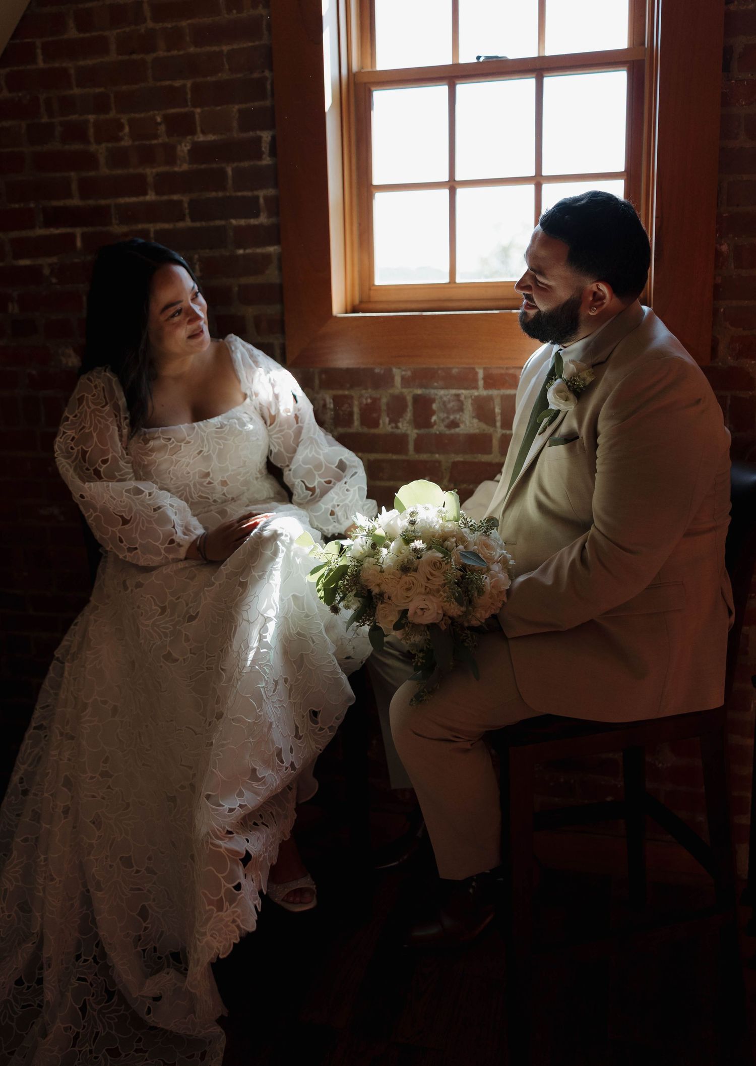 Couple in formal attire embraces near a sunlit window in an industrial brick space.