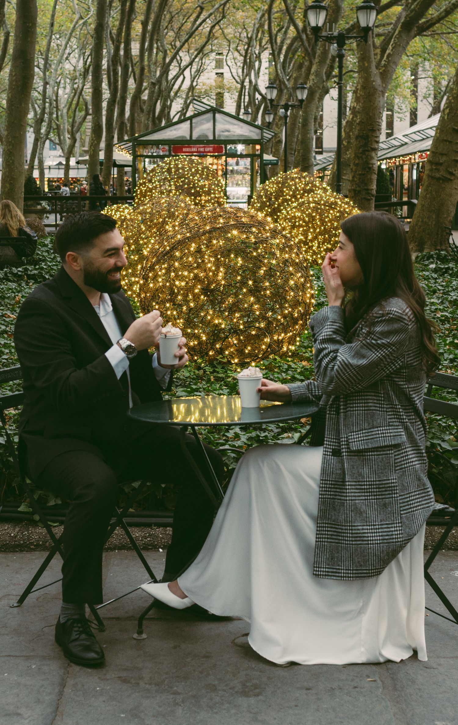 A couple drinks coffee at a cafe table with festive lights in the background.