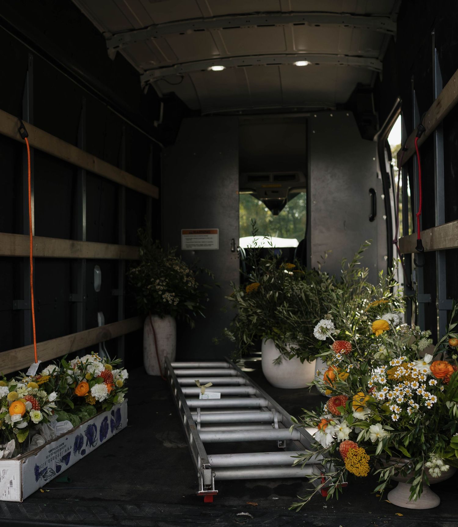 Inside a delivery truck filled with floral arrangements and ladders with natural light streaming through the back.