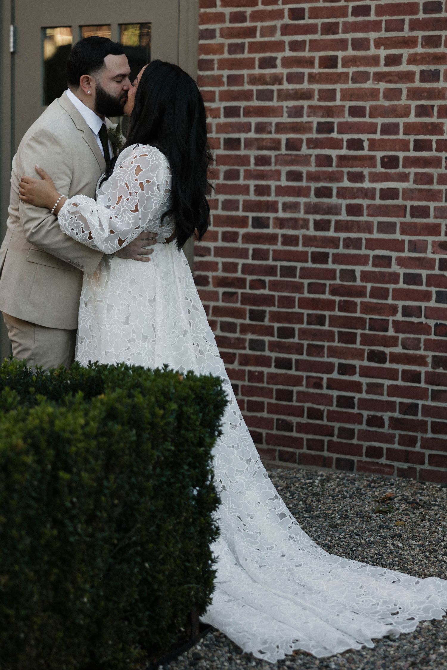 Couple in wedding attire embracing against a brick wall backdrop.