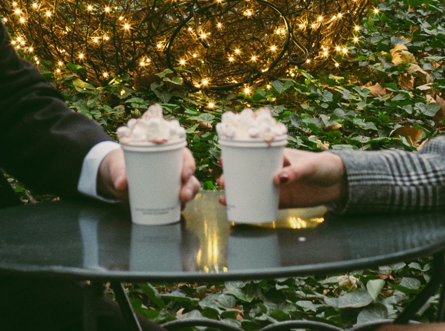 Two mugs of hot chocolate with whipped cream against a backdrop of twinkling lights.