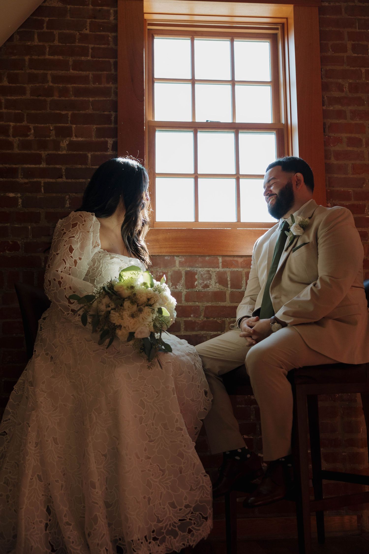 Wedding couple seated in rustic brick room with streaming window light.