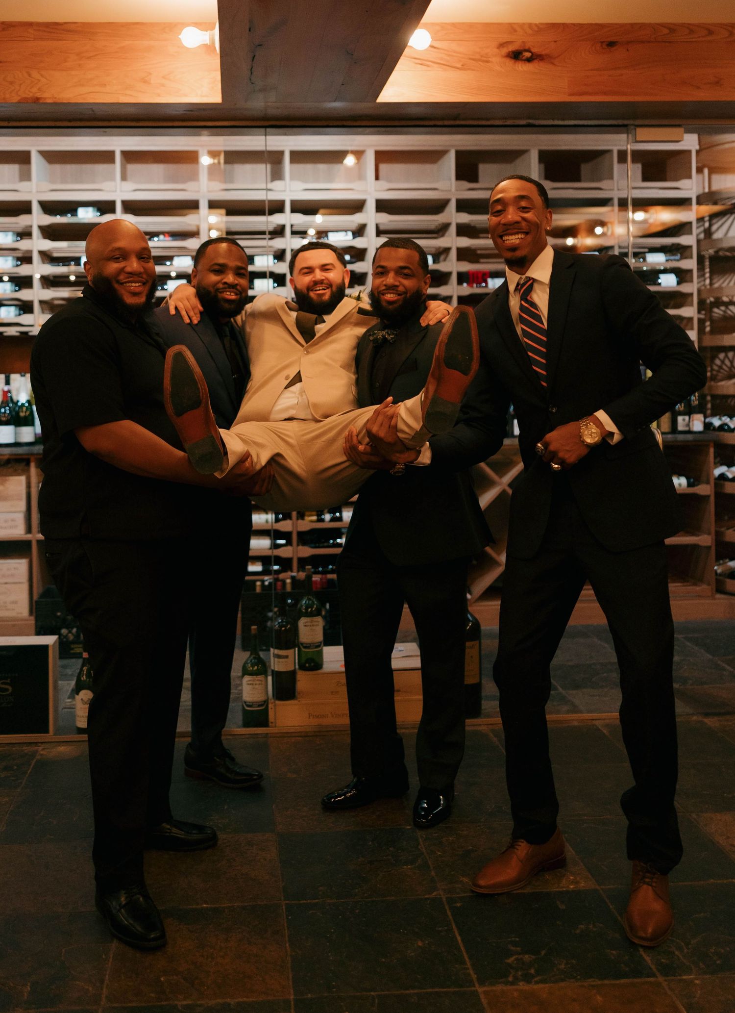 Groomsmen share a lighthearted moment in a wine cellar during a wedding celebration.