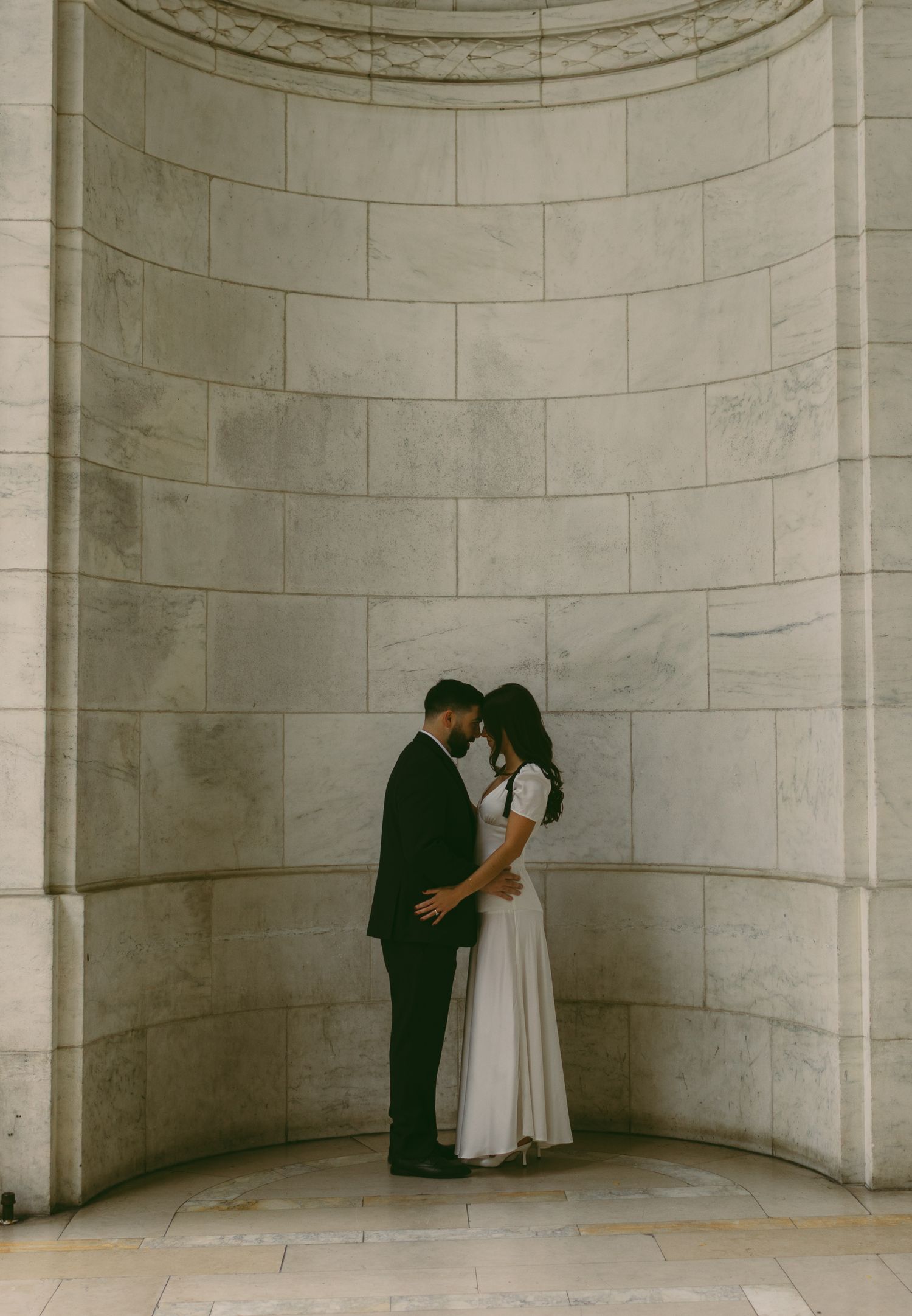 A couple shares an intimate moment in an alcove with classic marble architecture.