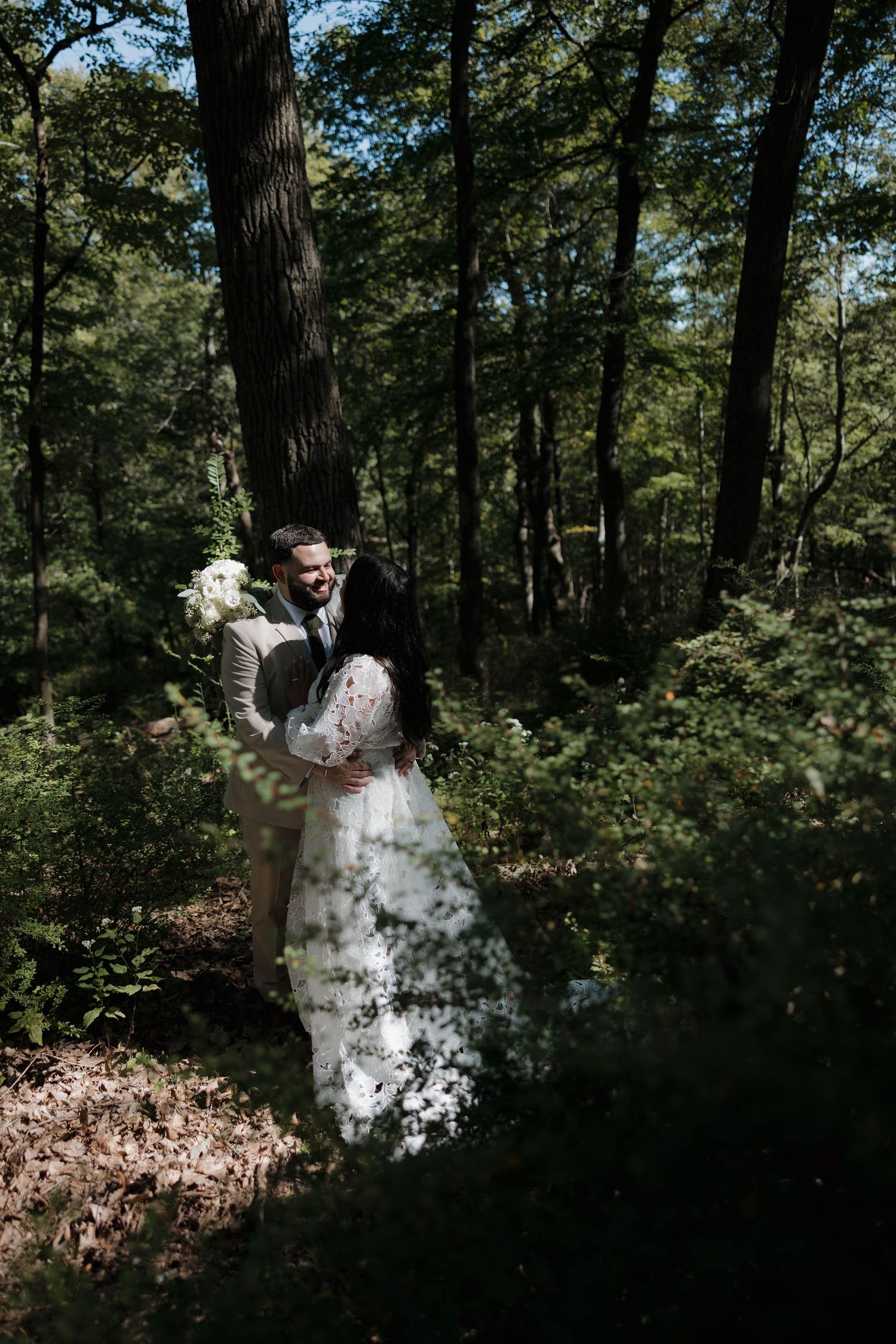 Newlyweds share a romantic dance among tall trees in a wooded setting with dappled sunlight.