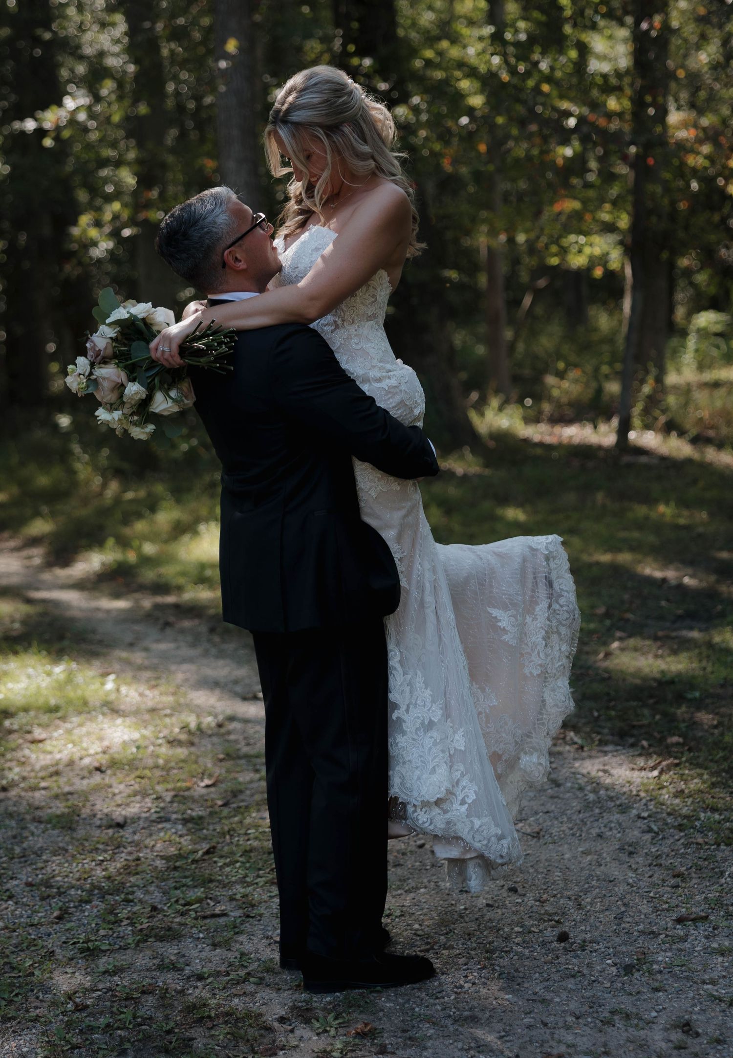 A romantic wedding lift pose captured in a sunlit wooded setting.