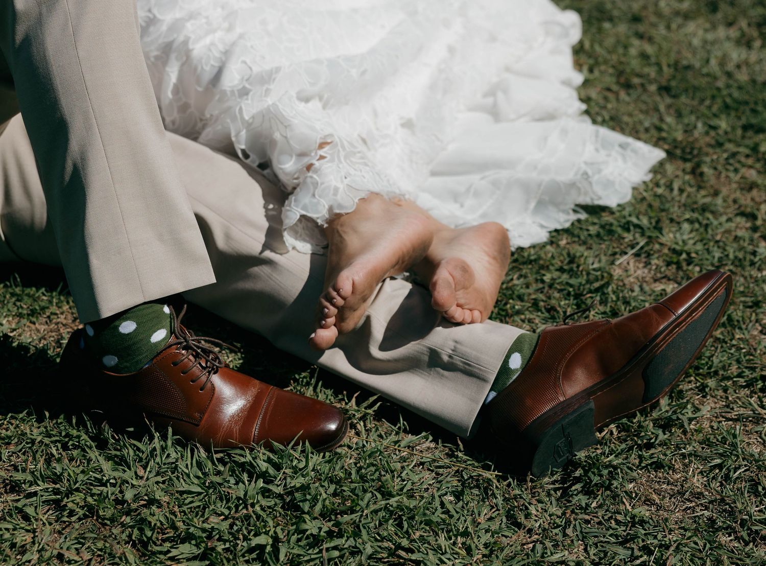 Brown leather dress shoes and white wedding dress hem visible on grass during outdoor wedding.