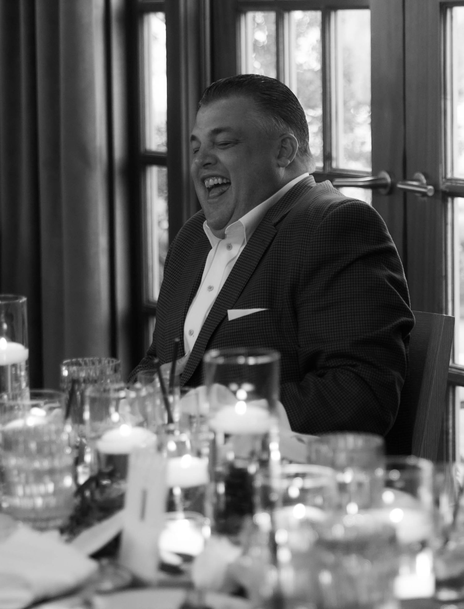 A black and white candid photo of laughter during a wedding reception with glasses on the table.