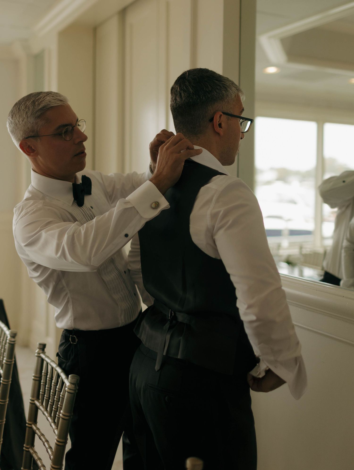 Men in formal attire help each other prepare in a bright indoor setting.