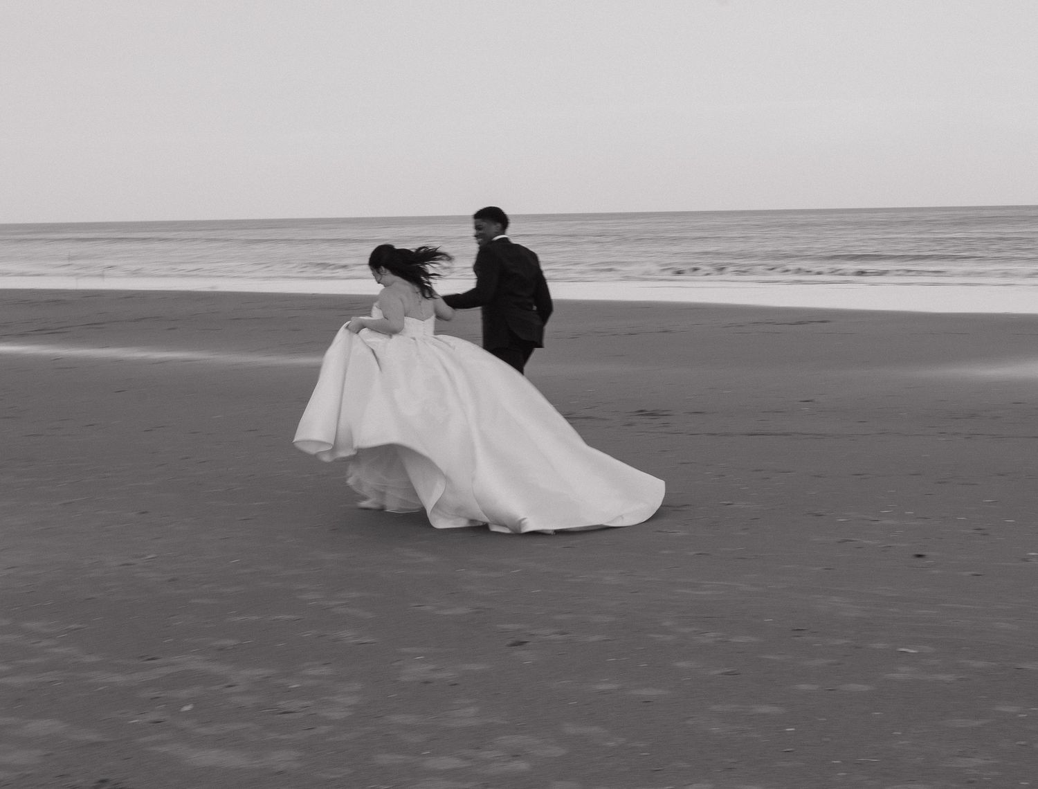 A couple in wedding attire walks along an empty beach in black and white photography.