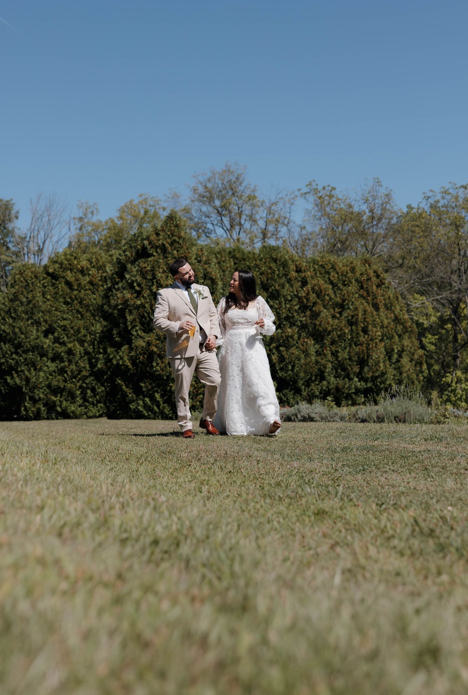Wedding couple sitting together on grassy hillside with bushes in background.