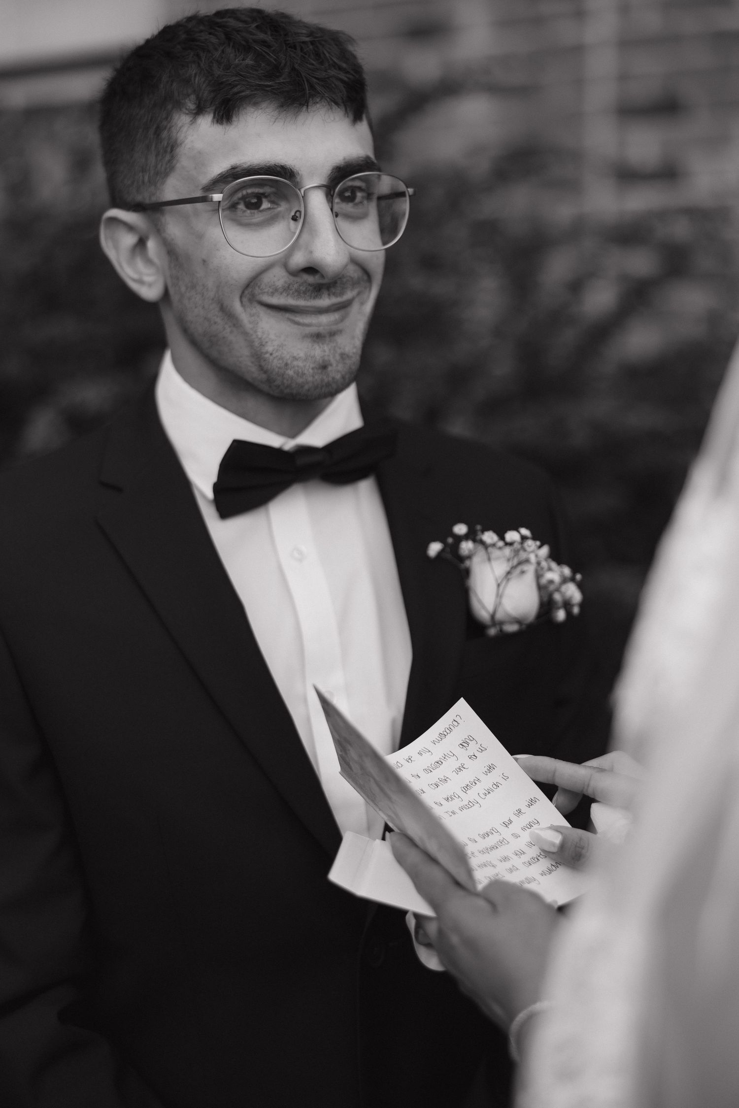 A formal black and white portrait of someone in a tuxedo reading a handwritten note.